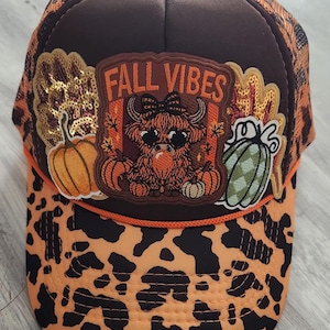 May include: A brown and orange leopard print trucker hat featuring a central patch with the text FALL VIBES, a highland cow illustration, and pumpkin graphics. The design includes sequined accents and a bright orange cord trim across the brim.