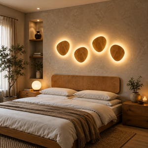May include: Four organic-shaped wooden wall sconces mounted above a bed, each featuring a warm, glowing backlight. The light fixtures have a smooth, light-brown wood grain finish and are arranged in a horizontal row against a textured wall.