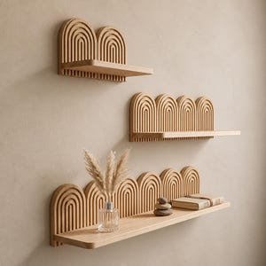 Puede incluir: Tres estantes de pared de madera de color claro con paneles traseros arqueados y listonados. El estante superior tiene dos arcos, el del medio tiene cuatro y el inferior tiene cinco. Un jarrón de cristal con hierba seca, tres piedras apiladas y un libro descansan en el estante inferior.