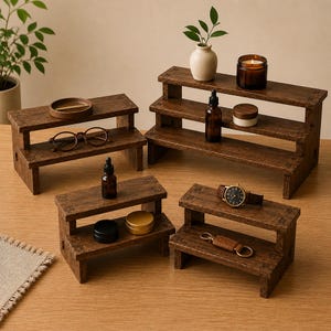 Peut inclure: Un ensemble de quatre présentoirs à étages en bois brun foncé de hauteurs et largeurs variées. Les supports présentent une conception en gradins avec des surfaces planes pour organiser de petits objets comme des flacons en verre ambré.