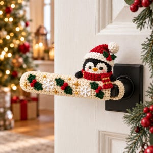 Op de afbeelding: Een crèmekleurige gehaakte deurkrukhoes met een pinguïn die een rood-witte kerstmuts en sjaal draagt. De hoes is versierd met geborduurde groene hulstblaadjes, rode bessen, witte sneeuwvlokken en kleine gouden sterretjes.