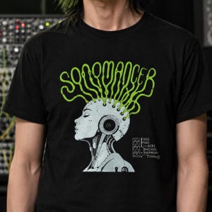May include: A black t-shirt featuring a graphic of a grey cyborg head in profile with green, flowing, vine-like lines extending from the top. The text reads "SONOMANCER" in green and "FREQ MIND MINE MACHINE HARMONY" in white with wave icons.