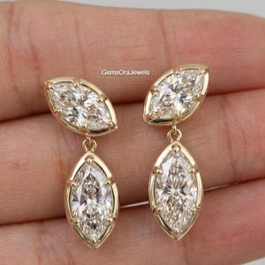 May include: A pair of drop earrings featuring two marquise-cut clear stones set in gold-toned metal. Each earring consists of a smaller marquise stone at the top and a larger marquise stone suspended below. The text GemsOraJewels is visible.