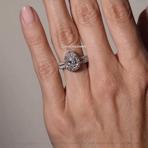 May include: A silver-toned ring set featuring a pear-shaped center stone surrounded by a halo of smaller round stones, with a matching pavé band. The text GemsOraJewels is visible above the ring, which is worn on a finger.