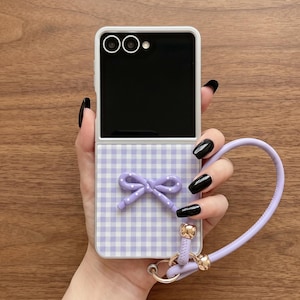 May include: A foldable smartphone case featuring a light purple and white gingham pattern. The case includes a three-dimensional purple bow accent in the centre and a matching light purple wrist strap attached with gold-toned metal hardware.