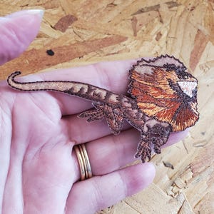 May include: Embroidered patch of a frilled-neck lizard. The patch features detailed stitching in shades of brown, tan, and orange, with a prominent frill around the head. The lizard's tail is curved, and its legs are visible.