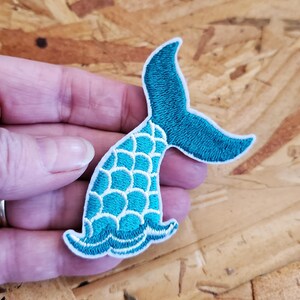 May include: Embroidered mermaid tail patch in teal and white. The tail fin is teal, and the body of the tail has a scale pattern in teal with white outlines. The patch has a white border.