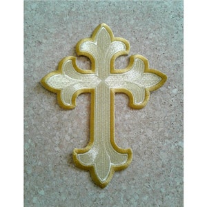 May include: Embroidered gold cross with ornate detailing. The cross features a light gold fill with a darker gold outline. The design is symmetrical and has a classic religious theme. The cross is likely a patch or applique.