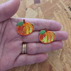 May include: Two embroidered pumpkin patches with orange and yellow sequins, green leaves, and brown stems. The patches are small, round, and likely for decorative purposes, possibly for fall-themed crafts or apparel.
