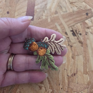 May include: Embroidered patch featuring two acorns with teal caps and orange bodies, accompanied by a tan oak leaf and green pine needles. The patch is held in a hand, showcasing the detailed stitching and autumnal theme.