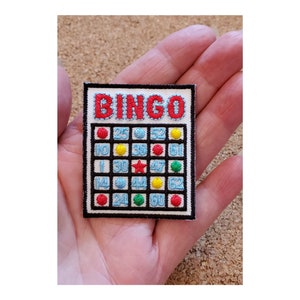 May include: A black and white embroidered patch with the word "BINGO" in red letters. The patch features a traditional bingo card with numbers and colored dots.
