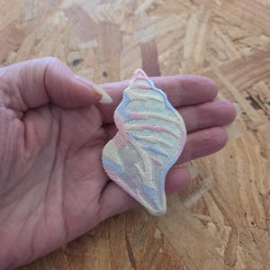 May include: An embroidered patch in the shape of a seashell, featuring a pastel color palette of light blue, soft pink, and pale yellow threads. The design includes intricate stitching that mimics the natural ridges and spiral form of a shell.