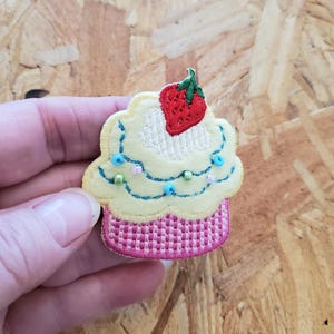 May include: A yellow and pink embroidered cupcake with white frosting and a red strawberry on top. The cupcake is decorated with blue and green beads.