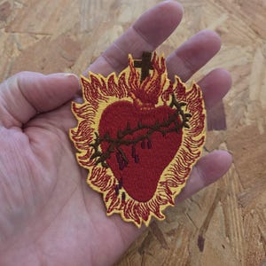 May include: An embroidered patch featuring a red heart encircled by a brown crown of thorns, topped with a small brown cross and surrounded by yellow and orange flames. The design is stitched with detailed thread work on a fabric base.
