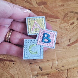 May include: Three embroidered alphabet blocks, each with a different letter: A, B, and C. The blocks are square, with a light blue border and a different color for each letter: pink, blue, and green. The background is a wood grain.