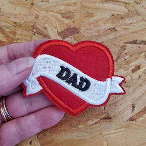 May include: A red heart-shaped patch with a white banner embroidered with the word "DAD" in black. The patch has a red border and is likely made of felt or a similar fabric. This patch is a decorative item.