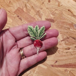 May include: Embroidered radish patch with a vibrant red root and green leaves. The radish has a detailed, textured appearance, showcasing the intricate stitching. This handmade patch is a unique accessory for clothing or crafts.