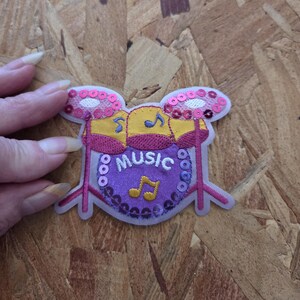May include: An embroidered iron-on patch shaped like a drum set. The design features a purple bass drum with the word MUSIC and a yellow musical note embroidered in white and yellow thread. It is accented with pink and purple sequins.