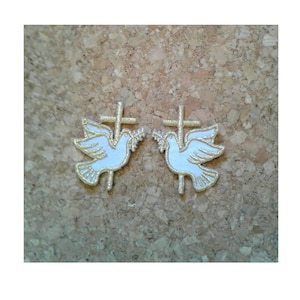 May include: Two white doves with gold trim holding olive branches in their beaks, each perched on a gold cross. The doves and crosses are embroidered on a white fabric.