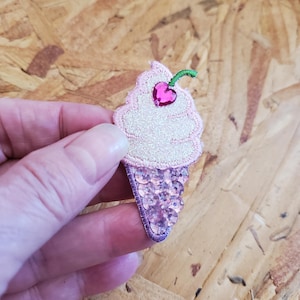 May include: A pink and white ice cream cone patch with a sequined cone and a pink cherry on top. The ice cream has a glittery texture and is outlined in pink thread. The cherry has a green stem.