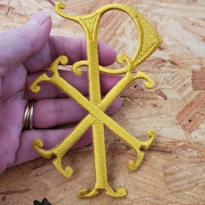 May include: A gold embroidered Chi Rho symbol, a monogram of Christ, featuring the Greek letters Chi (Χ) and Rho (Ρ). The symbol is intricately stitched with a textured appearance, and the design includes decorative flourishes.