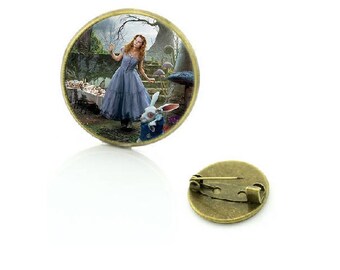 May include: A round, bronze-colored pin with a glass dome featuring a scene from Alice in Wonderland. The image shows Alice in a blue dress, standing in a forest with a white rabbit. The pin has a safety clasp on the back.