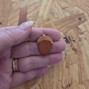 May include: An acorn-shaped patch with brown and tan embroidery. The patch features a detailed design with a textured appearance, showcasing the acorn's cap and body. This patch is suitable for adding to clothing or accessories.