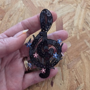May include: A treble clef patch covered in black sequins, featuring small embroidered light blue and pink star-shaped flowers along the edges. The musical symbol is crafted with a dark outline and detailed stitching, creating a textured appearance.
