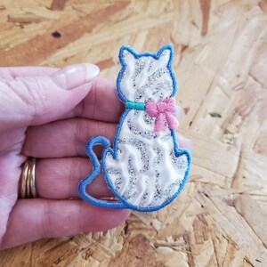 May include: A white cat-shaped patch with silver glitter accents, a blue outline, and a pink bow. The cat has a turquoise collar. The patch is designed to be sewn or ironed onto fabric. The cat is sitting.