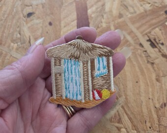 May include: An embroidered patch featuring a beach hut with a thatched roof, a light blue door, and a window. The design includes a small red and white striped ball and a yellow ball on the sand at the base of the structure, rendered in tan, blue, and red thread.