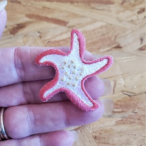 May include: A starfish-shaped embroidered patch featuring a white center with small gold-colored accents and a vibrant pink stitched border. The design is flat and detailed with a textured thread finish, showcasing a five-pointed marine creature shape.