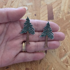 May include: Two embroidered patches in the shape of evergreen trees. The trees have green foliage and brown trunks. The patches are held in a hand, and the background is a light brown wood.
