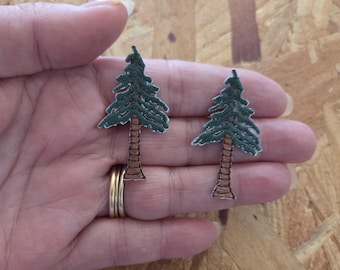 May include: Two embroidered patches shaped like evergreen trees. The trees have green foliage and brown trunks. The patches are held in a hand, with a light brown wood background.