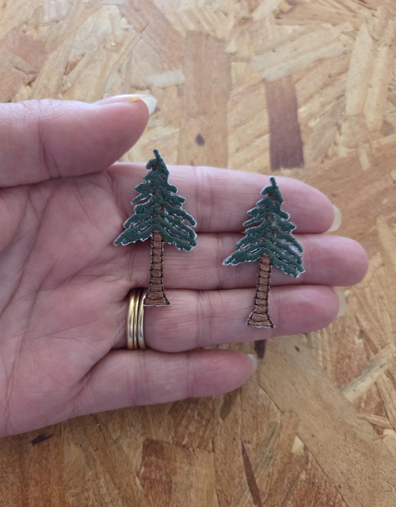 May include: Two embroidered patches in the shape of evergreen trees. The trees have green foliage and brown trunks. The patches are held in a hand, and the background is a light brown wood.