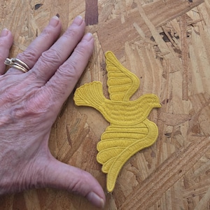May include: A mustard yellow embroidered patch in the shape of a bird in flight with wings spread. The patch features detailed stitching throughout the body and wings, showcasing a textured surface and defined edges for the avian silhouette.