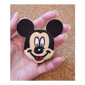 May include: A brown and black embroidered patch of Mickey Mouse's face with a red smile.