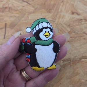 May include: Embroidered penguin patch wearing a green hat and scarf. The penguin has a white belly, black body, and yellow feet. The scarf has red, blue, and green stripes. The patch is designed for sewing or ironing onto fabric.