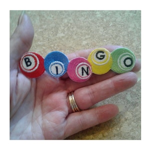 May include: A set of six colorful felt bingo balls with the letters B, I, N, G, and O embroidered on them.