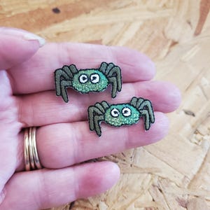 May include: Two embroidered spider patches. Each patch features a green body with black legs and white eyes. The patches are small, measuring approximately 1 inch in width, and are designed for sewing or ironing onto fabric.