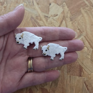 May include: Two white embroidered bison patches with tan horns and hooves are displayed. The patches are on a hand, and the bison are in different sizes. The patches are likely for sewing onto clothing or accessories.