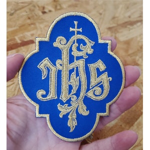 May include: A blue and gold embroidered patch with the letters "IHS" in a stylized font. The letters are surrounded by a decorative floral design and a cross at the top.