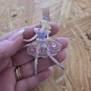 May include: An embroidered patch of a ballerina figure featuring a purple bodice, blonde hair, and a sheer white tulle skirt embellished with four round pink sequins. The figure is depicted in a dance pose with slender limbs and ballet slippers.