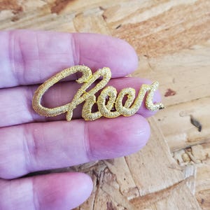 May include: A gold-colored embroidered patch with the word "Cheer" in a cursive font. The patch is held in a person's hand, and the background is a wooden surface.