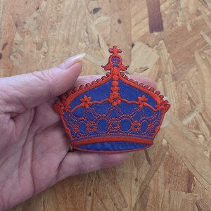 May include: An embroidered patch in the shape of a crown, featuring a vibrant blue base fabric with intricate orange thread detailing. The design includes a central cross at the top, decorative floral accents, and a scalloped edge along the crown.