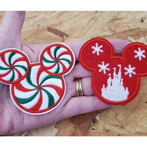 May include: Two red and white embroidered iron-on patches shaped like Mickey Mouse ears. One patch features a peppermint swirl design and the other features a white silhouette of Cinderella's castle with snowflakes.