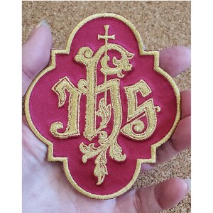May include: A red embroidered patch with a gold "IHS" monogram in a decorative font. The monogram is surrounded by a floral design and a cross at the top.