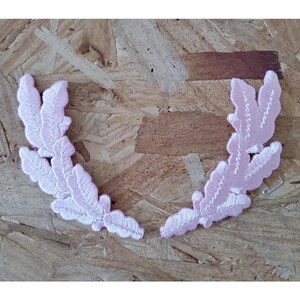 May include: Two light pink embroidered leaf-like appliques. Each applique has a detailed, feathery design with white stitching. The appliques are arranged in a semi-circular shape, suggesting they could be used as decorative elements for clothing or crafts.