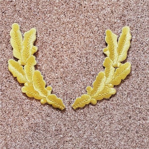 May include: Two gold embroidered laurel wreaths, each with multiple leaves, are arranged in a U-shape on a brown corkboard background.