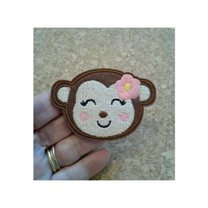 May include: Embroidered patch of a smiling monkey with a pink flower on its head. The monkey has brown fur, white eyes, and pink cheeks.