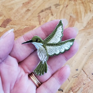 May include: Embroidered hummingbird patch in shades of green, white, and black. The patch features detailed stitching, showcasing the bird's wings, body, and beak. The hummingbird is depicted in mid-flight, with its wings spread. This patch is suitable for clothing or accessories.